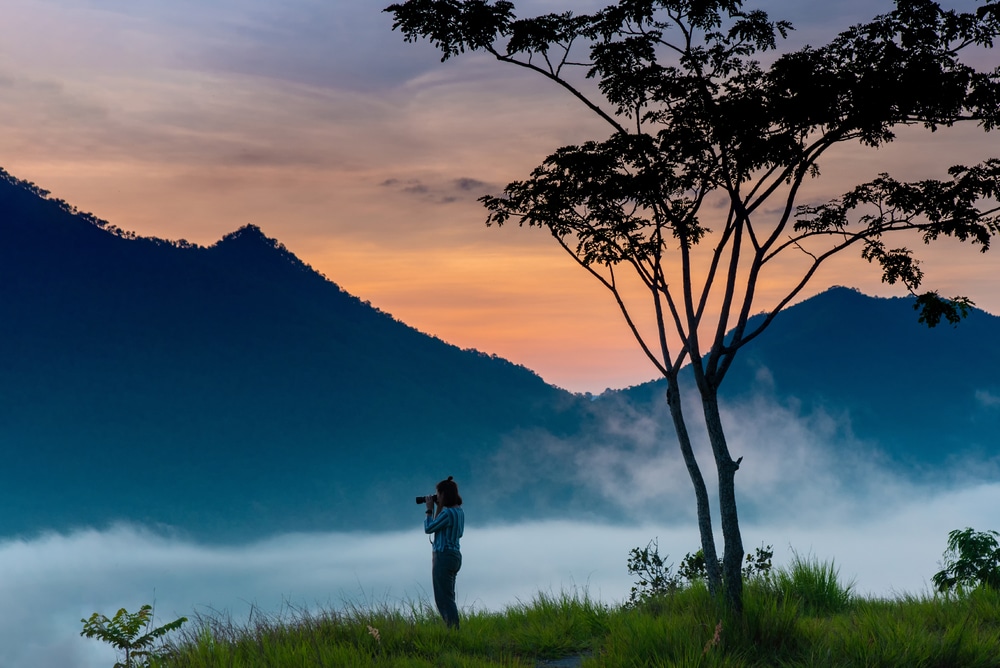ภาพคนกำลังถ่ายภาพทิวทัศน์ภูเขาที่ปกคลุมด้วยหมอกในช่วงรุ่งอรุณหรือพลบค่ำ โดยมีต้นไม้อยู่ด้านหน้า และเมฆลอยอยู่ใต้ยอดเขา — ฉากที่ถ่ายทอดความงามอันสงบของที่เที่ยวเชียงคาน สำหรับผู้ที่กำลังมองหาการผจญภัยที่น่าจดจำ.