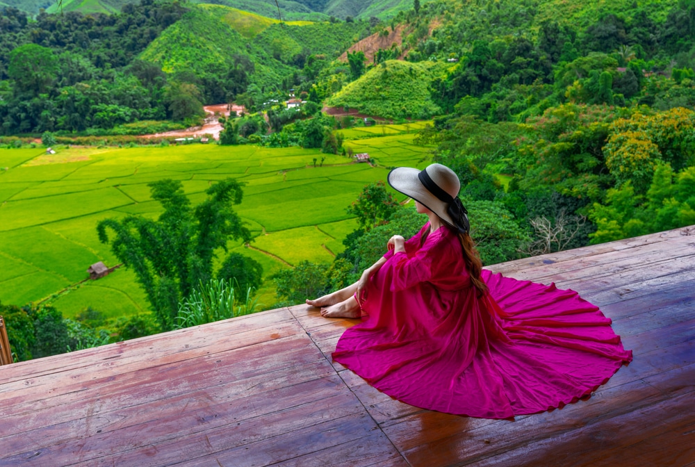 มีคนหนึ่งสวมหมวกปีกกว้างและชุดสีแดงนั่งอยู่บนระเบียงไม้ที่มองออกไปเห็นทุ่งนาสีเขียวและภูเขา สะท้อนความงามอันสงบของ น่านที่เที่ยว ซึ่งเป็นหนึ่งในจุดท่องเที่ยวที่ไม่ควรพลาดสำหรับผู้ที่ต้องการ เที่ยวน่าน.