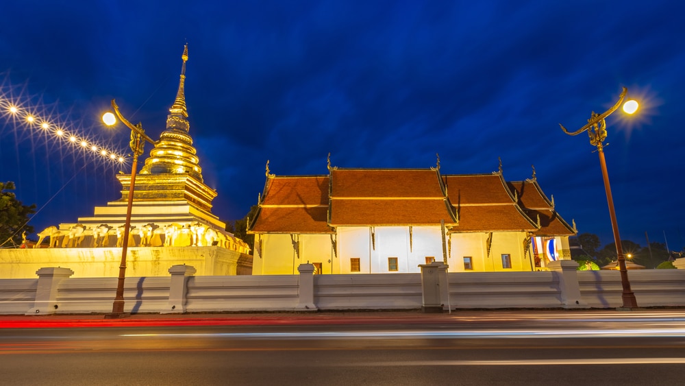วัดพุทธที่สว่างไสวและเจดีย์สีทองตั้งอยู่เบื้องหลังกำแพงสีขาวในยามค่ำคืน พร้อมด้วยแสงไฟถนนและแสงไฟรถยนต์ที่พร่ามัวในเบื้องหน้า — เป็นภาพที่ชวนหลงใหล ซึ่งถ่ายทอดความงามอันสงบของสถานที่ท่องเที่ยวในน่าน สำหรับผู้ที่ต้องการเที่ยวน่าน.