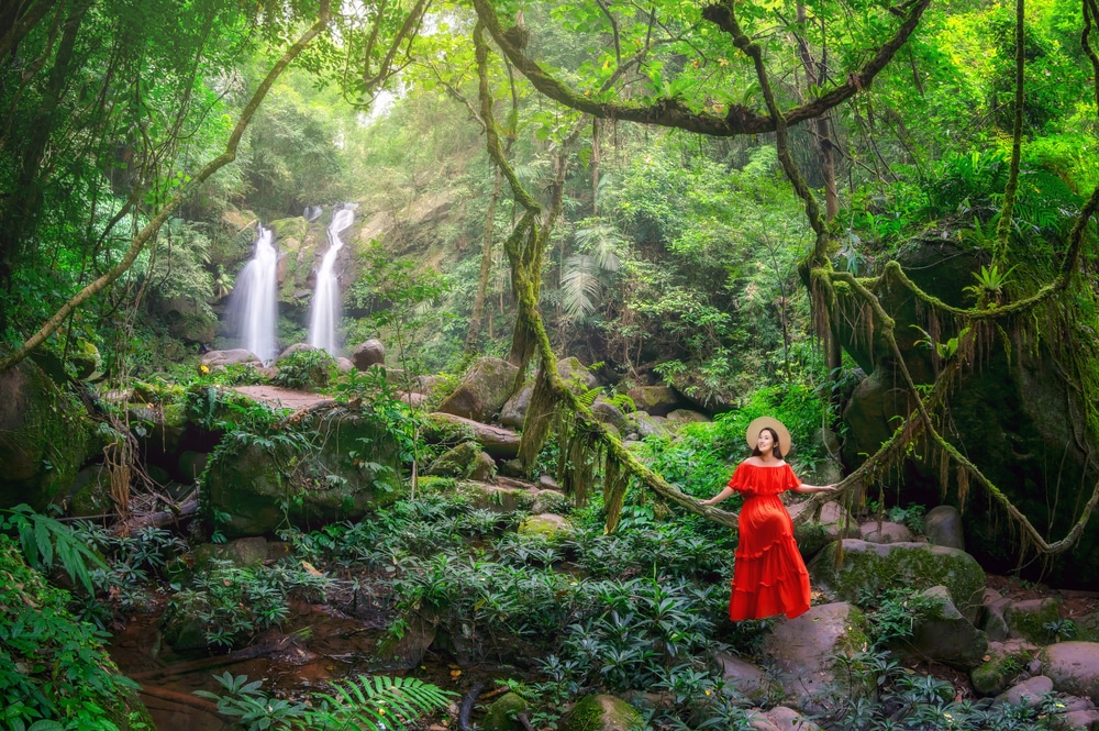 ผู้หญิงคนหนึ่งสวมชุดสีแดงและหมวก กำลังยืนอยู่บนท่อนไม้ที่ปกคลุมด้วยมอส ในป่าเขียวชอุ่มที่มีน้ำตกสองแห่งเป็นฉากหลัง สะท้อนความงามอันสงบของ น่านที่เที่ยว—หนึ่งในสถานที่ท่องเที่ยวน่านที่น่าหลงใหลที่สุดสำหรับผู้ที่รักธรรมชาติ.
