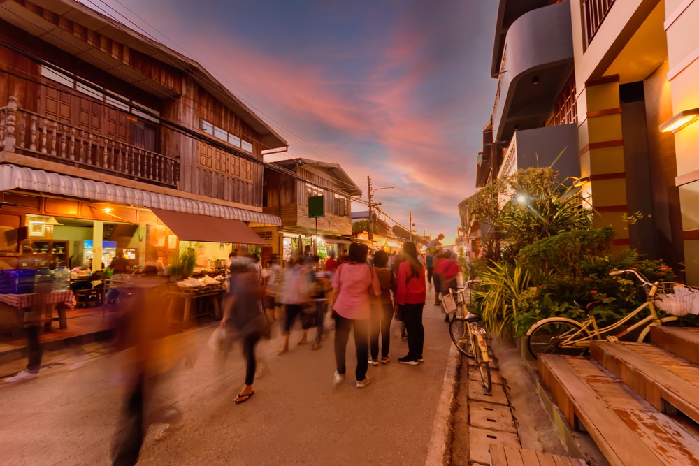 ภาพตลาดถนนที่คึกคักยามพลบค่ำในเชียงคานที่เที่ยว โดยมีผู้คนเดินผ่านไปมาอย่างเบลอๆ ข้างอาคารไม้ที่น่ารัก ร้านอาหารที่คึกคัก แสงไฟที่อบอุ่น และจักรยานสีเหลืองที่จอดอยู่ — ถ่ายทอดบรรยากาศที่ชวนให้มาเที่ยว ซึ่งดึงดูดผู้คนจำนวนมากให้มาสำรวจและเที่ยวเชียงคาน.