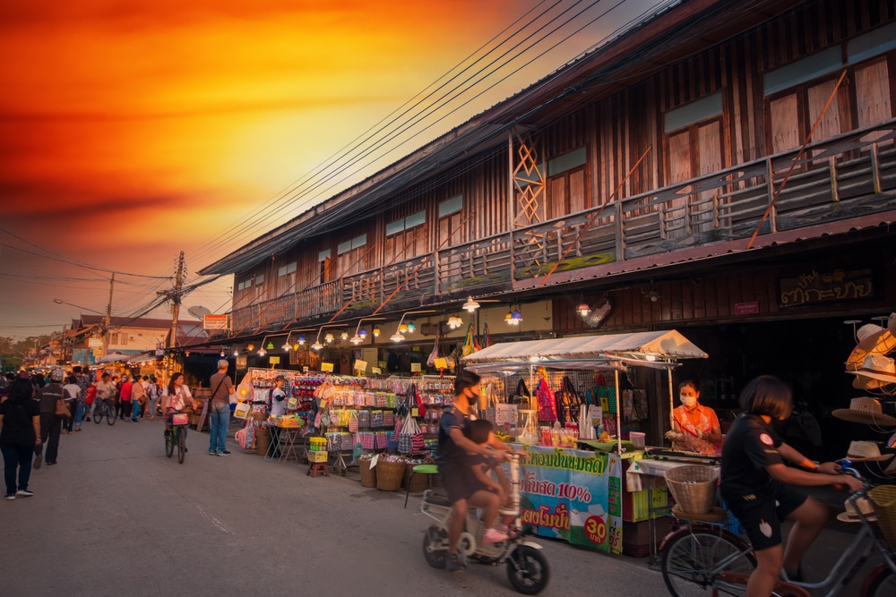 ตลาดกลางแจ้งที่มีหน้าร้านทำจากไม้ ผู้ขายสินค้า และผู้ซื้อเดินหรือขี่จักรยานใต้ท้องฟ้าสีส้มยามพระอาทิตย์ตกดิน — เป็นภาพที่คลาสสิกเมื่อคุณมาเที่ยวที่เที่ยวเชียงคาน.
