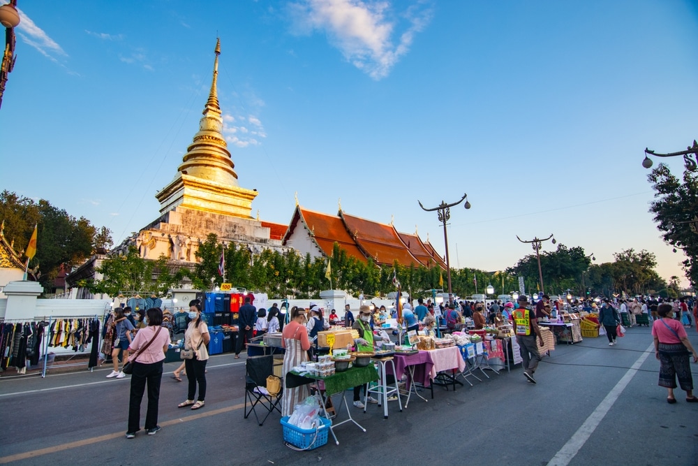 ผู้คนเดินชมร้านค้าในตลาดกลางแจ้งที่ตั้งอยู่ริมถนนข้างวัดใหญ่ที่มียอดเจดีย์สูงตระหง่าน ท่ามกลางท้องฟ้าสีครามสดใส ซึ่งทำให้ผู้มาเยือนได้สัมผัสวัฒนธรรมท้องถิ่นที่เต็มไปด้วยชีวิตชีวาและบรรยากาศอันเป็นเอกลักษณ์ที่ทำให้การเที่ยวน่านมีเสน่ห์ไม่เหมือนใคร.