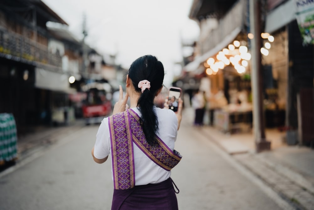 ผู้หญิงคนหนึ่งสวมผ้าคาดเอวลายดอกยืนอยู่บนถนนและถ่ายภาพด้วยโทรศัพท์มือถือ เพื่อบันทึกบรรยากาศที่เต็มไปด้วยสีสันของการท่องเที่ยวเชียงคาน ในพื้นหลังสามารถเห็นร้านค้าและแสงไฟได้ ซึ่งสะท้อนถึงเสน่ห์ที่คึกคักของจุดท่องเที่ยวยอดนิยมแห่งนี้ในเชียงคาน.