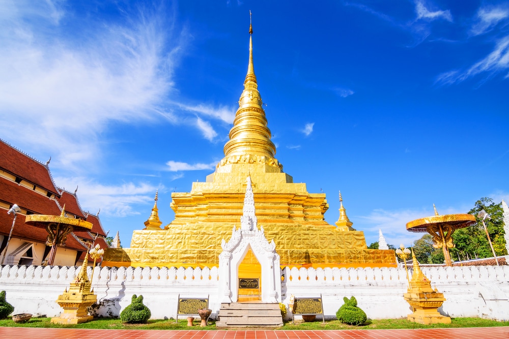 เจดีย์สีทองที่มีรายละเอียดประดับตกแต่งอย่างวิจิตรตั้งอยู่ใต้ท้องฟ้าสีคราม ล้อมรอบด้วยกำแพงสีขาวและพืชพรรณเขียวขจีภายในบริเวณวัด — เป็นหนึ่งในสถานที่ท่องเที่ยวในน่านที่ไม่ควรพลาด สำหรับผู้ที่ต้องการสัมผัสความงามทางวัฒนธรรมของการท่องเที่ยวน่าน.