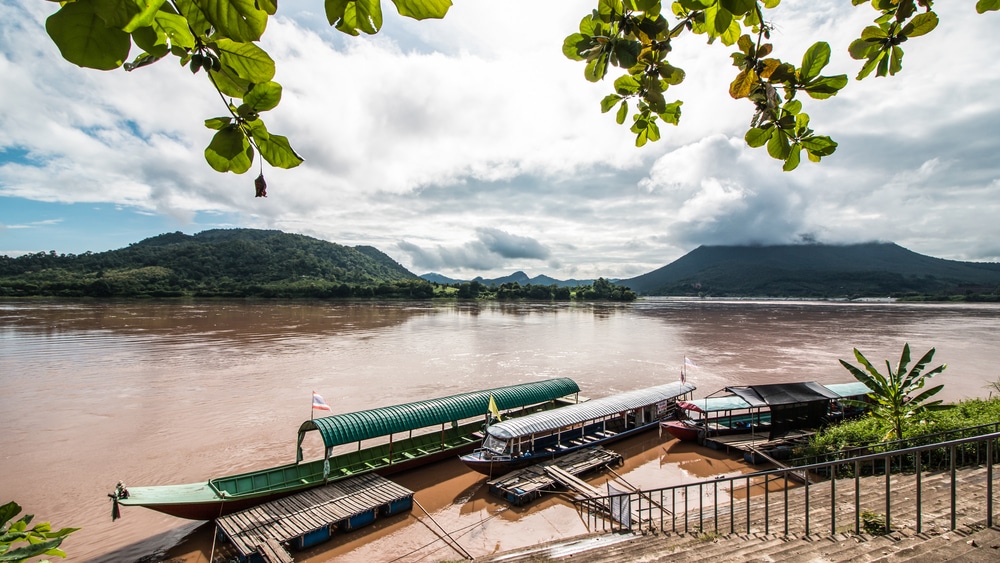 เรือยาวสามลำจอดอยู่ริมแม่น้ำสายกว้าง โดยมีภูเขาสีเขียวและท้องฟ้าครึ้มเป็นฉากหลัง — ภาพที่สงบเงียบนี้สะท้อนความเสน่ห์ของเที่ยวเชียงคานได้อย่างสมบูรณ์แบบ ซึ่งเป็นหนึ่งในสถานที่ท่องเที่ยวที่ห้ามพลาดในเชียงคานสำหรับผู้ที่รักธรรมชาติ.