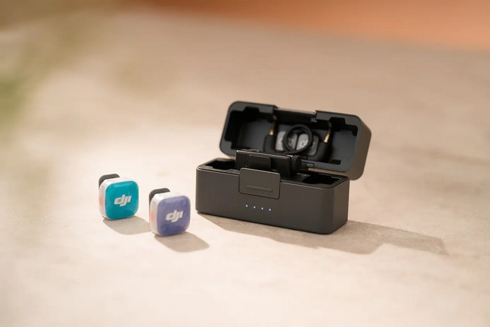 หูฟังไร้สายสองข้างที่มีโลโก้ “DJI” ถูกวางไว้ข้างกล่องชาร์จสีดำที่เปิดอยู่ บนพื้นผิวสีกลาง.