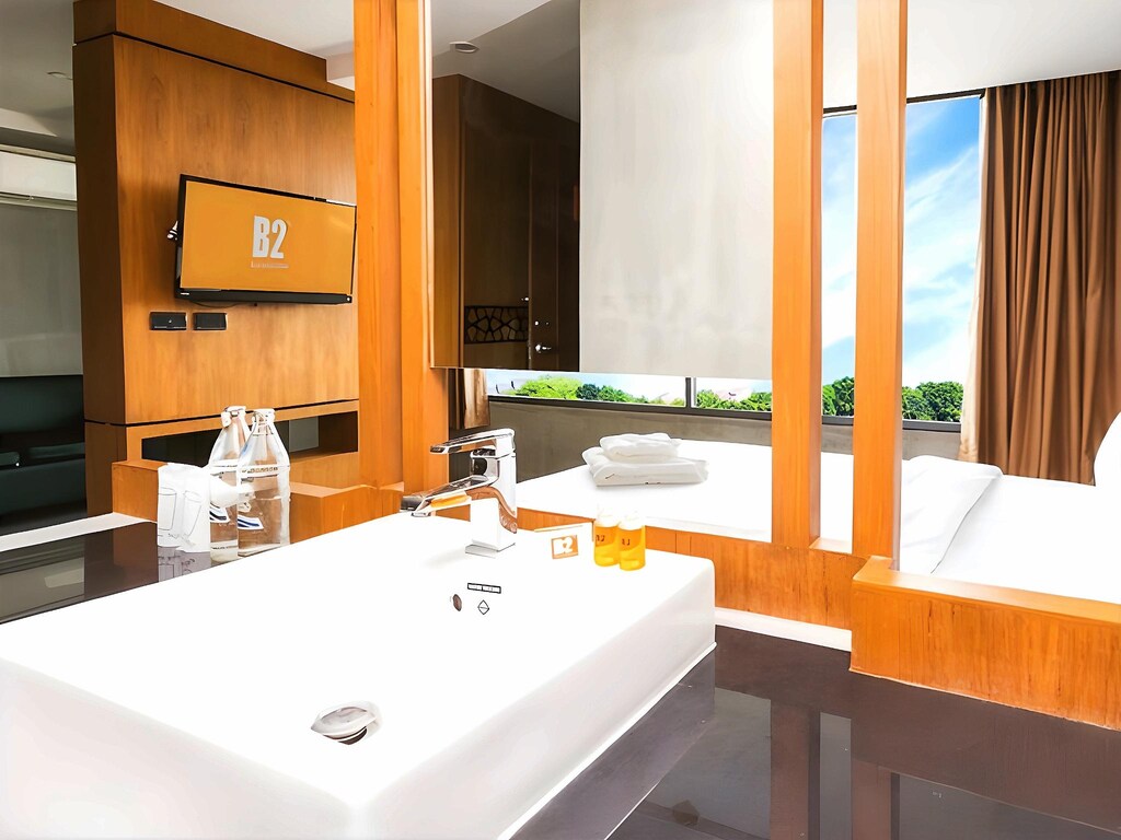 ห้องพักโรงแรมสมัยใหม่ที่มีอ่างล้างหน้าสีขาว ผลิตภัณฑ์ดูแลร่างกาย และขวดน้ำอยู่ด้านหน้า ส่วนด้านหลังมีเตียงนอน โทรทัศน์ กระจกขนาดใหญ่ และหน้าต่างที่มองเห็นวิวสวยงาม — เหมาะอย่างยิ่งสำหรับผู้ที่กำลังมองหา โรงแรมขอนแก่น หรือ ที่พักขอนแก่น ที่ทันสมัยและสะดวกสบาย.
