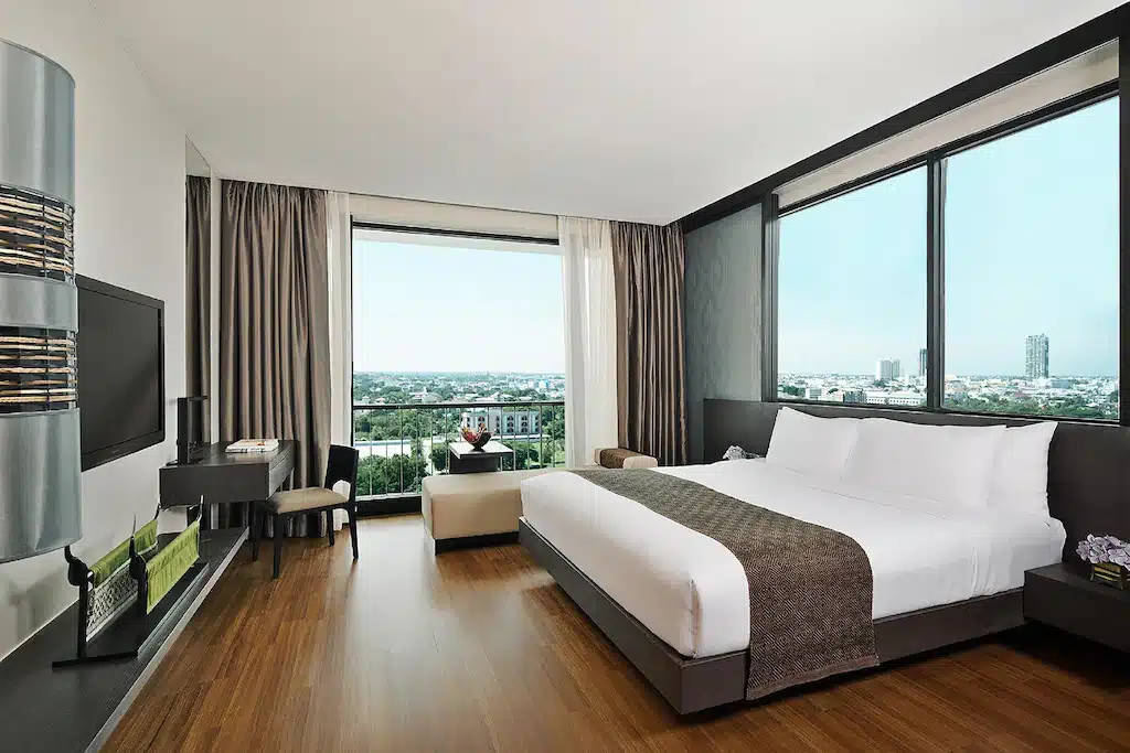 ห้องพักสไตล์โมเดิร์นในโรงแรมขอนแก่น พร้อมด้วยเตียงขนาดใหญ่ พื้นไม้ โต๊ะทำงาน ทีวี และหน้าต่างสูงจากพื้นถึงเพดานที่ให้มุมมองเมืองอันงดงาม—เหมาะอย่างยิ่งสำหรับการพักของคุณในขอนแก่น.