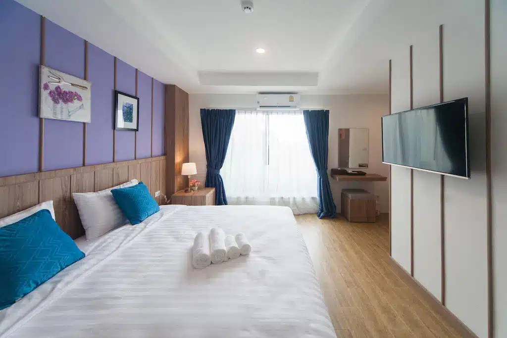 ห้องพักโรงแรมสไตล์โมเดิร์น พร้อมเตียงขนาดใหญ่ หมอนสีฟ้า ผ้าเช็ดตัวม้วนเป็นรูปทรง TV ติดผนัง พื้นไม้ โต๊ะทำงาน โคมไฟ และหน้าต่างขนาดใหญ่พร้อมผ้าม่านสีฟ้า — เหมาะอย่างยิ่งสำหรับผู้ที่กำลังมองหาที่พักขอนแก่นหรือโรงแรมในขอนแก่นที่มีสไตล์ สำหรับการพักในเมืองนี้.