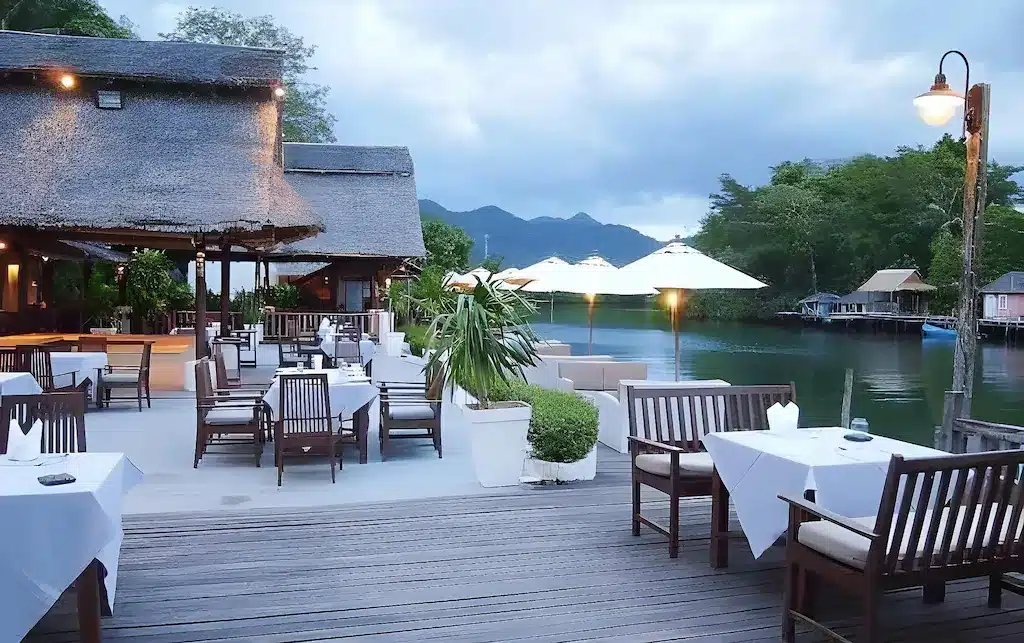 ร้านอาหารกลางแจ้งริมแม่น้ำ พร้อมโต๊ะและเก้าอี้ไม้ ผ้าปูโต๊ะสีขาว ร่มขนาดใหญ่ และพืชพรรณเขียวชอุ่ม ท่ามกลางฉากหลังของภูเขาและน้ำนิ่งสงบใต้ท้องฟ้าที่มีเมฆครึ้ม — เป็นประสบการณ์การรับประทานอาหารที่สมบูรณ์แบบสำหรับแขกที่กำลังมองหา ที่พักเกาะช้าง หรือ โรงแรมเกาะช้าง ในบรรยากาศธรรมชาติที่สงบเงียบ.