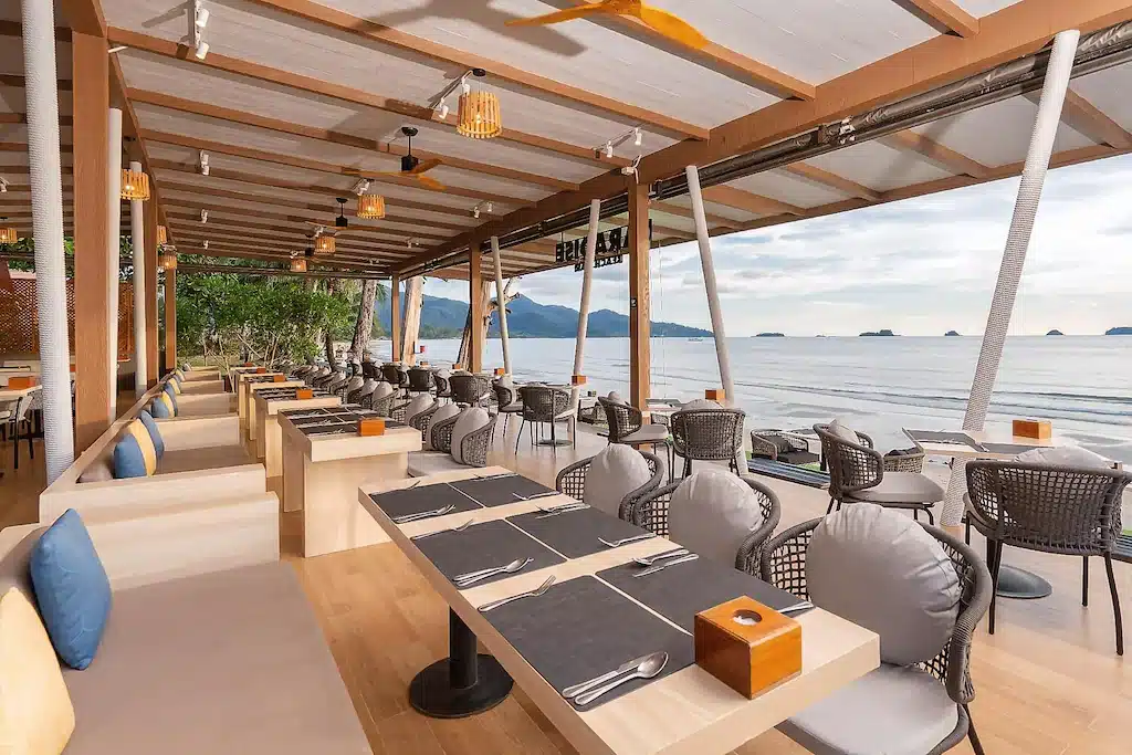 ร้านอาหารกลางแจ้งสมัยใหม่ที่มีโต๊ะจัดวางอย่างเป็นระเบียบและเก้าอี้มีเบาะนั่ง พร้อมวิวชายหาดที่สงบและทะเลกว้างใหญ่ พร้อมเกาะๆ ที่อยู่ไกลๆ ใต้ท้องฟ้าที่มีเมฆบางส่วน—เป็นสถานที่ที่สมบูรณ์แบบสำหรับแขกที่กำลังมองหาที่พักเกาะช้างหรือโรงแรมเกาะช้าง และชื่นชอบการรับประทานอาหารอย่างหรูหราริมทะเล.