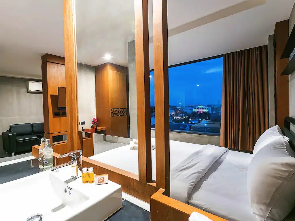 ห้องพักโรงแรมสไตล์ทันสมัย พร้อมเตียงขนาดใหญ่ อ่างล้างหน้า และพื้นที่นั่งเล่น; มีหน้าต่างสูงจากพื้นถึงเพดานที่มองเห็นวิวเมืองยามพลบค่ำ มอบประสบการณ์การพักที่สะดวกสบายในหนึ่งในโรงแรมที่ดีที่สุดของขอนแก่น.