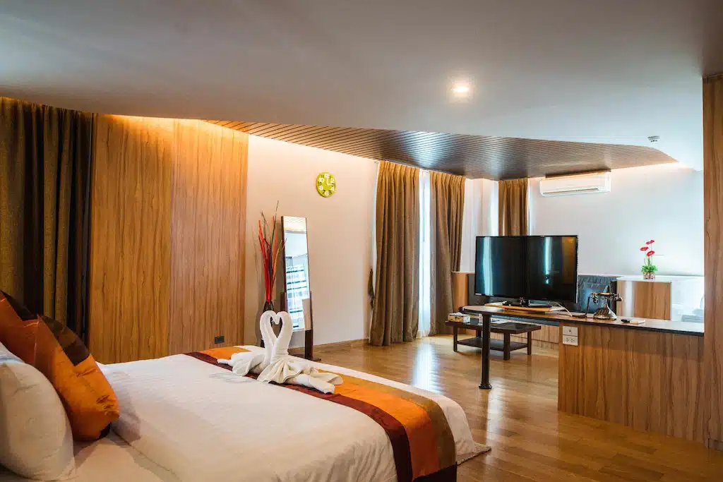 ห้องพักโรงแรมสไตล์ทันสมัย พร้อมเตียงคู่ ทีวี เฟอร์นิเจอร์ไม้ หน้าต่างขนาดใหญ่พร้อมผ้าม่าน ผ้าเช็ดตัวตกแต่งที่พับเป็นรูปหงส์ และโต๊ะเล็กพร้อมเก้าอี้ — เหมาะอย่างยิ่งสำหรับแขกที่ต้องการการพักผ่อนที่สะดวกสบายที่โรงแรมในขอนแก่น.