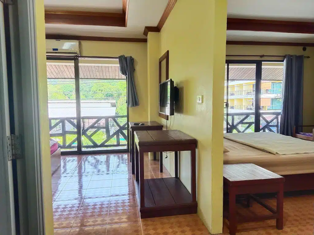ห้องพักในโรงแรมที่เต็มไปด้วยแสงแดด มีพื้นปูกระเบื้อง เตียงนอน โทรทัศน์ติดผนัง และหน้าต่างขนาดใหญ่พร้อมม่าน มองออกสู่ระเบียงและพื้นที่สีเขียว เหมาะอย่างยิ่งสำหรับการเข้าพักที่ โรงแรมเกาะช้าง หรือสำหรับใครก็ตามที่กำลังมองหา เกาะช้างที่พัก ที่ผสมผสานความสะดวกสบายกับวิวทิวทัศน์ที่สวยงาม.