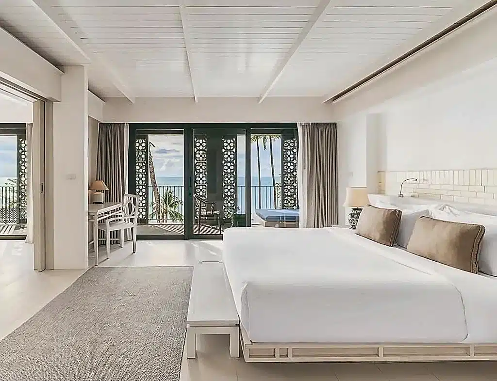 ห้องพักโรงแรมสมัยใหม่ที่มีเตียงขนาดใหญ่ โต๊ะทำงาน และระเบียงที่มองเห็นวิวทะเล ตกแต่งด้วยโทนสีกลางๆ และประตูเลื่อนลายสวย — เหมาะสำหรับผู้ที่กำลังมองหา โรงแรมเกาะช้าง ที่ผสมผสานความสบายและสไตล์เข้ากับวิวทะเลที่สวยงาม.