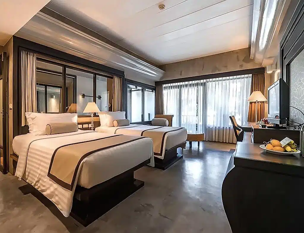 ห้องพักโรงแรมสมัยใหม่ที่มีเตียงคู่สองเตียง โต๊ะทำงานพร้อมโคมไฟ ทีวีจอแบน ชามผลไม้ หน้าต่างขนาดใหญ่พร้อมม่าน และการตกแต่งด้วยสีโทนกลาง — เหมาะอย่างยิ่งสำหรับแขกที่ต้องการความสะดวกสบายอย่างมีสไตล์ที่ โรงแรมเกาะช้าง.