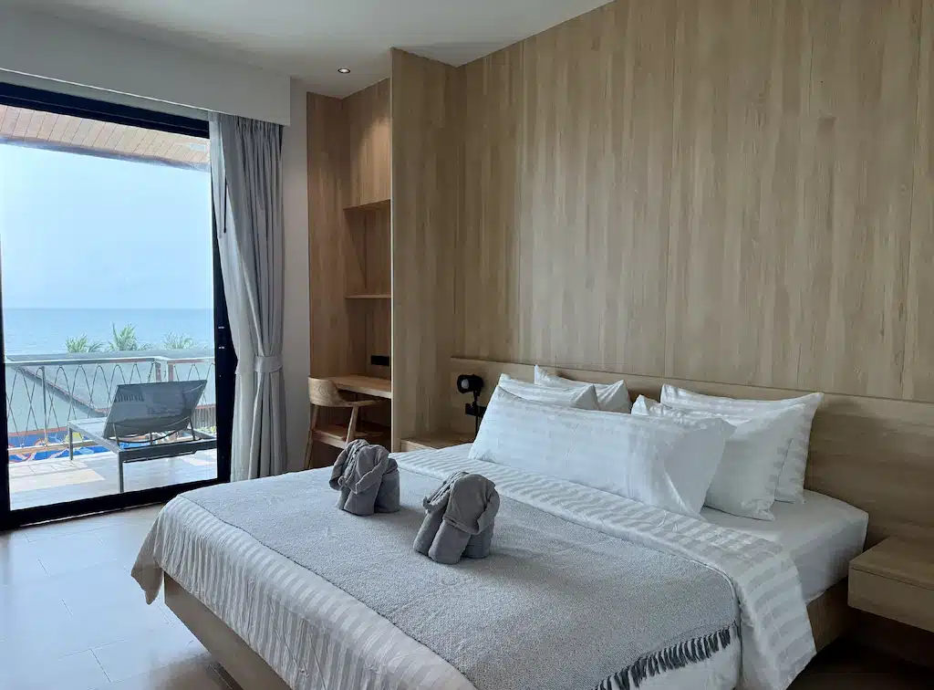 ห้องพักโรงแรมสมัยใหม่ที่มีเตียงจัดแต่งอย่างเรียบร้อย สัตว์ทำจากผ้าขนหนูสองตัว เฟอร์นิเจอร์ไม้สีอ่อน โต๊ะทำงาน และหน้าต่างขนาดใหญ่พร้อมระเบียงที่มองเห็นวิวทะเล — เหมาะอย่างยิ่งสำหรับผู้ที่มองหาที่พักบนเกาะช้างที่มีสไตล์และสะดวกสบาย.