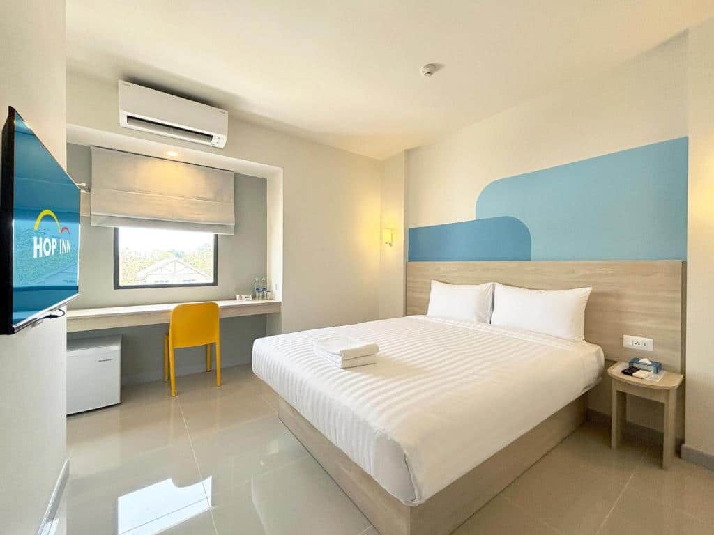 ห้องพักสมัยใหม่ที่โรงแรมในขอนแก่น มีเตียงคู่ที่จัดแต่งอย่างเรียบร้อย โต๊ะทำงานพร้อมเก้าอี้สีเหลือง ทีวีติดผนัง ตู้เย็นขนาดเล็ก และหน้าต่างที่มีม่านบังแสง ผ้าเช็ดตัวถูกวางไว้บนเตียงเพื่อความสะดวกของคุณ ทำให้ที่นี่เป็นตัวเลือกที่สมบูรณ์แบบสำหรับผู้ที่กำลังมองหาที่พักในขอนแก่น ที่ผสมผสานทั้งความสบายและสไตล์.