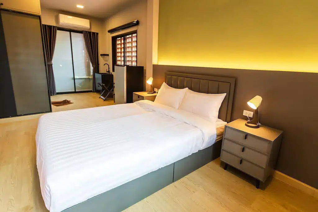 ห้องนอนสไตล์โมเดิร์นที่มีเตียงคู่ ชุดเครื่องนอนสีขาว โต๊ะข้างเตียงสองตัวพร้อมโคมไฟ พื้นไม้ และหน้าต่างขนาดใหญ่พร้อมผ้าม่าน — เหมาะอย่างยิ่งสำหรับผู้ที่กำลังมองหาที่พักขอนแก่น หรือต้องการประสบการณ์การพักที่โรงแรมขอนแก่นอย่างมีสไตล์.