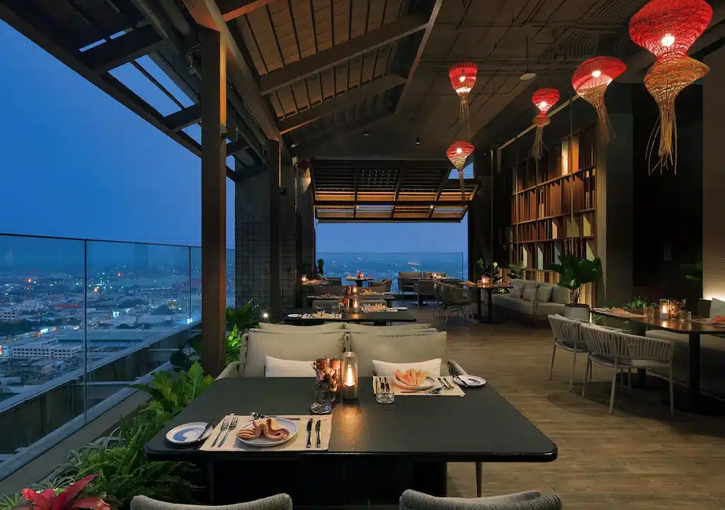 ร้านอาหารบนดาดฟ้าที่มีวิวเมืองยามพลบค่ำ พร้อมโต๊ะจัดไว้เรียบร้อย โคมไฟสีแดงแขวนประดับ และการตกแต่งสไตล์โมเดิร์นด้วยพืชพรรณและชั้นวางหนังสือ — เหมาะอย่างยิ่งสำหรับแขกที่ต้องการประสบการณ์การรับประทานอาหารอย่างมีสไตล์ที่ รีสอร์ทขอนแก่น หรือ โรงแรมขอนแก่น.