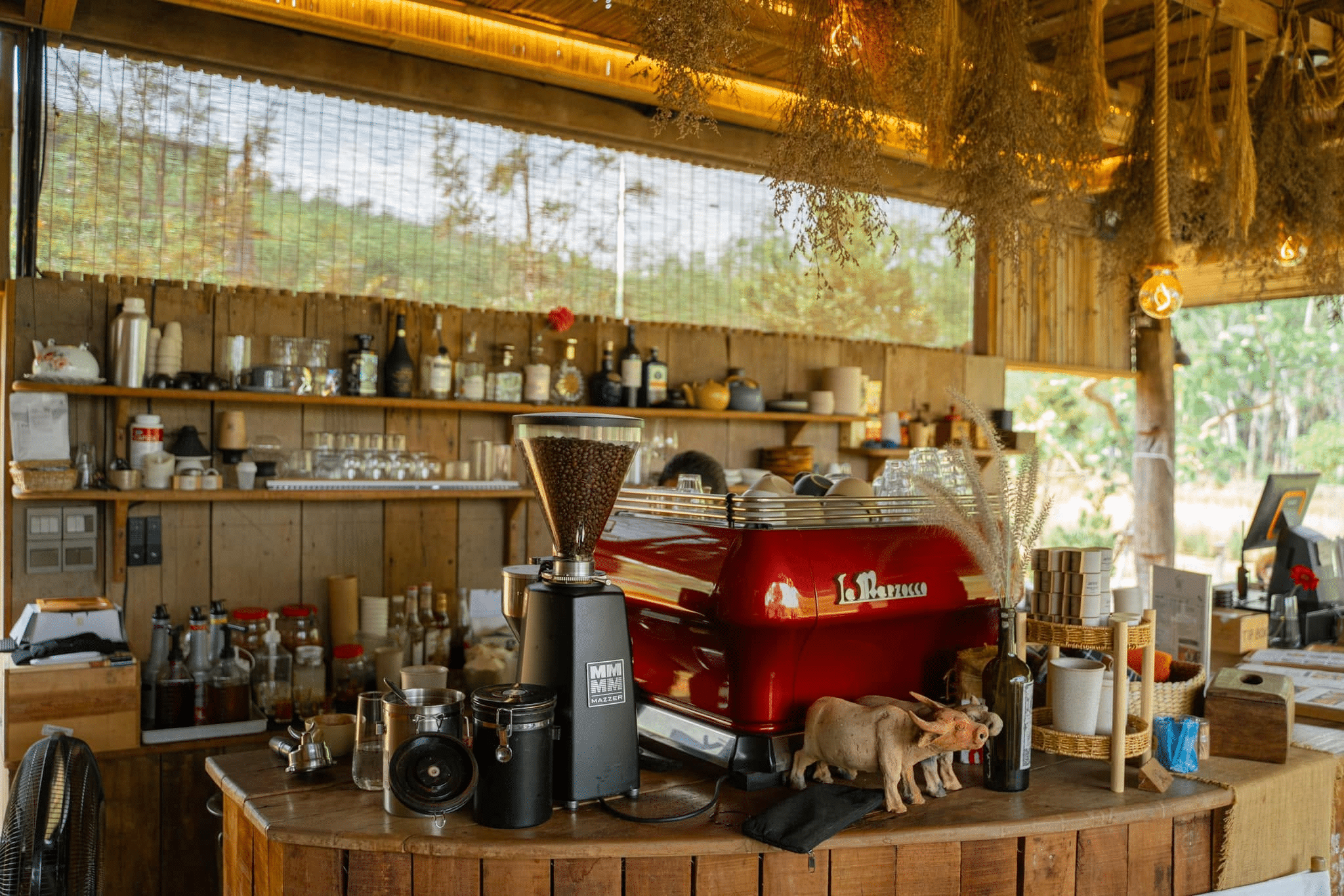 เคาน์เตอร์ร้านกาแฟที่มีเครื่องเอสเพรสโซสีแดง เครื่องบดกาแฟ ขวดแก้ว ถ้วย และของตกแต่งวางอยู่บนเคาน์เตอร์ไม้ — สะท้อนความอบอุ่นและเสน่ห์ที่มักพบเห็นในร้านกาแฟใกล้สถานที่ท่องเที่ยวระยอง — พร้อมด้วยชั้นวางของและเครื่องดื่มในพื้นหลัง.