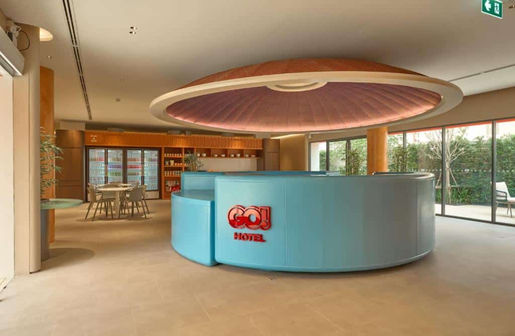 ล็อบบี้โรงแรมสมัยใหม่ที่มีเคาน์เตอร์รับแขกสีฟ้าอ่อนรูปโค้ง ป้าย “GO! HOTEL” สีแดง องค์ประกอบเพดานรูปวงกลม พื้นที่นั่งพักผ่อน และหน้าต่างสูงจากพื้นถึงเพดานที่มองเห็นวิวสวนด้านนอก — เป็นพื้นที่ที่ชวนให้เข้ามาพักผ่อน ซึ่งสะท้อนสไตล์สมัยใหม่ของโรงแรมขอนแก่นได้อย่างสมบูรณ์แบบ.