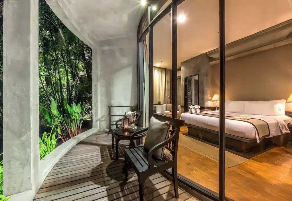 ห้องพักสไตล์ทันสมัยของโรงแรมเกาะช้าง ที่มีเตียงคิงไซส์ สามารถมองเห็นได้จากลานกลางแจ้งรูปโค้ง ซึ่งมีเก้าอี้ไม้สองตัวและโต๊ะหนึ่งตัว ล้อมรอบด้วยพืชพรรณเขียวชอุ่มในยามค่ำคืน.