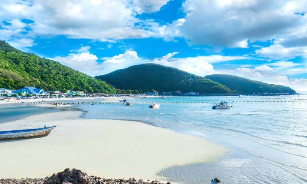 ชายหาดทรายขาวที่มีเรือเล็กในน้ำตื้น ภูเขาสีเขียวเป็นฉากหลัง และท้องฟ้าสีฟ้าที่มีเมฆบางส่วน—เป็นหนึ่งในสถานที่ท่องเที่ยวที่น่าหลงใหลของระยอง ที่มอบความสงบและผ่อนคลายท่ามกลางความงามของธรรมชาติ.