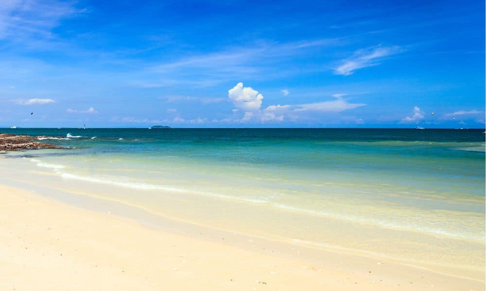 ชายหาดทรายขาวกับคลื่นเบาๆ น้ำทะเลสีฟ้าใส และท้องฟ้าที่มีเมฆบางส่วน มองเห็นเกาะเล็กๆ อยู่ไกลๆ ทำให้ที่นี่เป็นหนึ่งในสถานที่ท่องเที่ยวที่สวยงามที่สุดของระยอง สำหรับนักท่องเที่ยวที่มองหาความผ่อนคลายและความงามของธรรมชาติ.