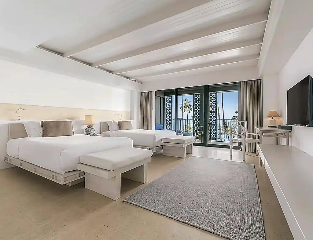 ห้องพักโรงแรมสไตล์ทันสมัยที่มีเตียงสองเตียง ม้านั่ง โต๊ะทำงาน โทรทัศน์ พรมขนาดใหญ่ และประตูบานเลื่อนกระจกที่เปิดออกสู่ระเบียงที่มีต้นปาล์มและวิวทะเล—เหมาะอย่างยิ่งสำหรับผู้ที่กำลังมองหา โรงแรมเกาะช้าง หรือ รีสอร์ทเกาะช้าง ที่ผสมผสานความสะดวกสบายกับทัศนียภาพเกาะที่สวยงามตระการตา.