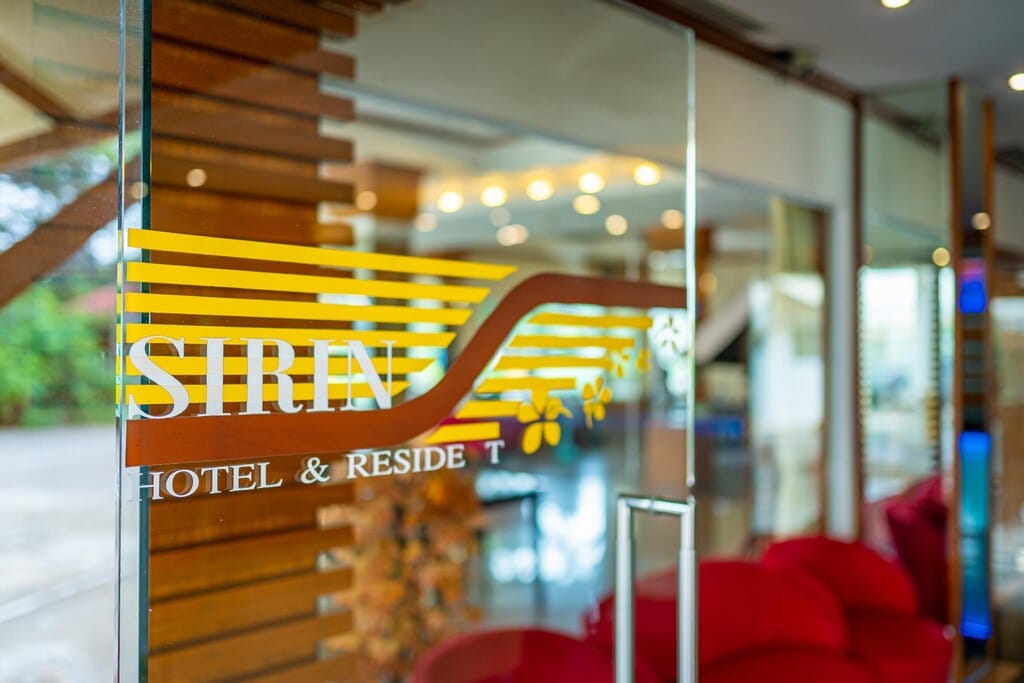 ประตูแก้วที่มีข้อความ “Sirin Hotel & Residence” พิมพ์อยู่ สะท้อนแสงและของตกแต่งภายในที่เบื้องหลัง ให้ภาพลักษณ์ที่อบอุ่นและน่าต้อนรับของหนึ่งในที่พักขอนแก่นหรือรีสอร์ทขอนแก่นที่โดดเด่น.