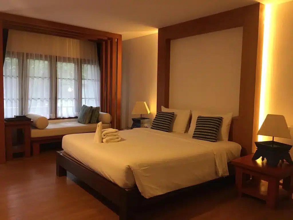ห้องพักในโรงแรมที่มีเตียงขนาดใหญ่ หมอนลายทาง โต๊ะข้างเตียงพร้อมโคมไฟ ที่นั่งริมหน้าต่าง พื้นไม้ และแสงไฟนุ่มนวล — เหมาะอย่างยิ่งสำหรับแขกที่กำลังมองหาโรงแรมในขอนแก่น เพื่อการพักผ่อนที่ผ่อนคลายและน่าจดจำ.