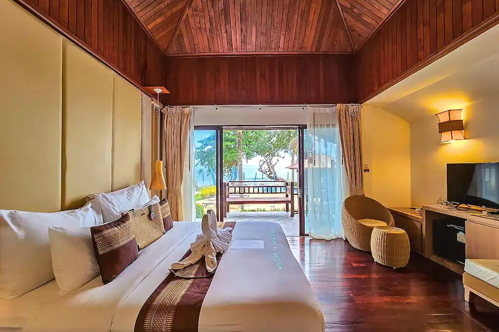 ห้องพักในโรงแรมที่มีเตียงขนาดใหญ่ เพดานไม้ ทีวี และพื้นที่นั่งเล่น พร้อมระเบียงที่มองเห็นวิวต้นไม้และทะเลผ่านประตูกระจกเลื่อน — เหมาะอย่างยิ่งสำหรับผู้ที่กำลังมองหาที่พักบนเกาะช้าง หรือโรงแรมบนเกาะช้าง ที่ล้อมรอบด้วยความงามของธรรมชาติ.