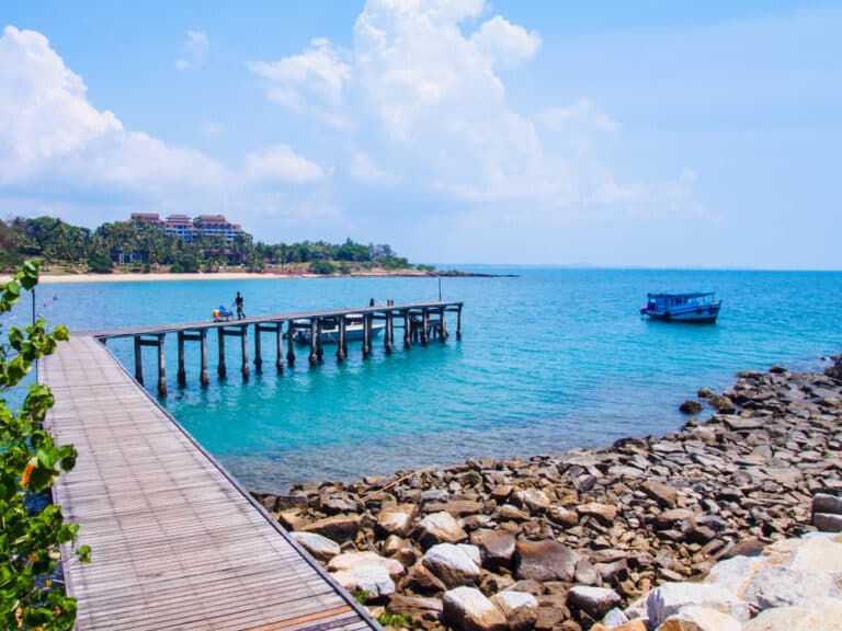 ท่าเรือไม้ที่ยื่นออกไปสู่ทะเลสีฟ้าใส พร้อมด้วยหินบนชายฝั่ง เรือลำหนึ่งที่ทอดสมออยู่ใกล้ๆ และผู้คนไม่กี่คนบนท่าเรือ ในพื้นหลังสามารถมองเห็นอาคารและต้นปาล์มได้ภายใต้ท้องฟ้าที่มีเมฆบางส่วน ฉากที่งดงามนี้ถือเป็นหนึ่งในสถานที่ท่องเที่ยวยอดนิยมของระยอง ทำให้เป็นจุดหมายที่ต้องไปเยือนสำหรับใครก็ตามที่กำลังมองหาสถานที่ท่องเที่ยวริมน้ำในระยอง.