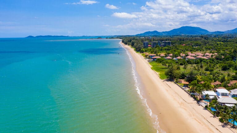 ภาพจากมุมสูงของชายหาดทรายที่มีน้ำสีเทอร์ควอยซ์ในระยอง ล้อมรอบด้วยพื้นที่สีเขียว อาคาร และภูเขาในระยะไกล ภายใต้ท้องฟ้าที่มีเมฆบางส่วน — เป็นตัวอย่างที่น่าทึ่งของสถานที่ท่องเที่ยวระยอง สำหรับใครก็ตามที่กำลังมองหาสถานที่ท่องเที่ยวระยองที่สวยงามดั่งภาพวาด.
