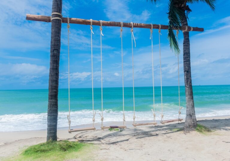ชิงช้าไม้ที่แขวนอยู่บนเชือกผูกระหว่างต้นปาล์มสองต้นบนชายหาดทราย พร้อมด้วยคลื่นทะเลสีเทอร์ควอยซ์และท้องฟ้าสีครามเป็นฉากหลัง — ภาพที่สงบและงดงาม ซึ่งสะท้อนเสน่ห์ของสถานที่ท่องเที่ยวระยอง สำหรับผู้ที่ต้องการพักผ่อนท่ามกลางความงามของธรรมชาติ.