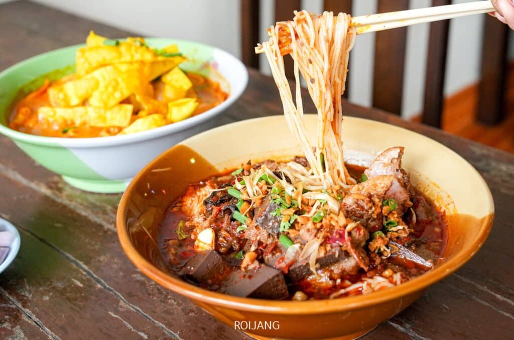 ก๋วยเตี๋ยวน้ำข้นรสเผ็ดใส่เนื้อสัตว์และผัก เคียงด้วยเต้าหู้ทอดในชามอีกใบ วางบนโต๊ะไม้ที่ ร้านอาหารเขาหลัก อย่างน่าเชื้อเชิญ ตะเกียบกำลังยกก๋วยเตี๋ยวรสเด็ดออกจากน้ำซุปร้อนๆ