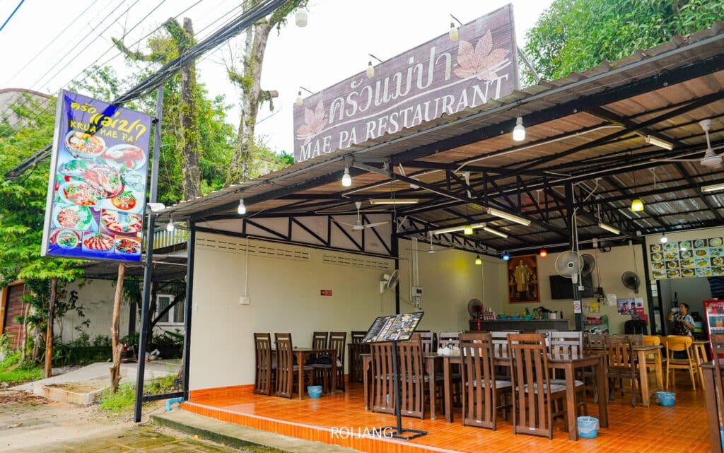 รูปภาพของร้านอาหารแม่ปะ ซึ่งเป็น ร้านอาหารเขาหลัก แสดงให้เห็นพื้นที่รับประทานอาหารแบบเปิดโล่ง มีโต๊ะและเก้าอี้ไม้ ป้ายพร้อมรูปภาพอาหารทางด้านซ้าย และป้ายร้านอาหารด้านบนทั้งภาษาไทยและภาษาอังกฤษ