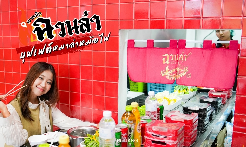 ผู้หญิงคนหนึ่งนั่งอยู่ที่โต๊ะในร้าน ร้านอาหารเขาหลัก ข้อความภาษาไทยอยู่ที่มุมขวาบน.อาหารกระเบื้องสีแดง ชื่อ ร้านอาหารเขาหลัก ถือตะเกียบและยิ้มอยู่บนโต๊ะ มีเครื่องปรุงและเครื่องดื่มต่างๆ วางอยู่ และมีสายพานลำเลียงอาหารอยู่ด้านหลัง.