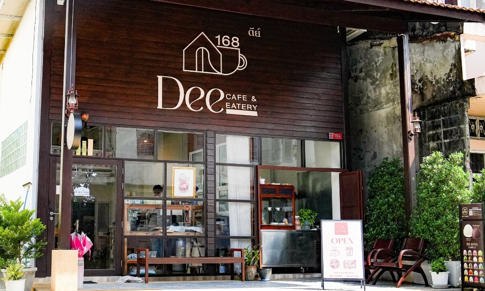 ด้านหน้าของ Dee Cafe & Eatery คาเฟ่บรรยากาศอบอุ่นที่ตกแต่งด้วยไม้และหน้าต่างบานใหญ่ ป้ายต้อนรับ "Open" เชิญชวนให้คุณเข้าไปด้านใน ต้นไม้และม้านั่งช่วยเพิ่มบรรยากาศให้ทางเข้าดูน่าดึงดูด.