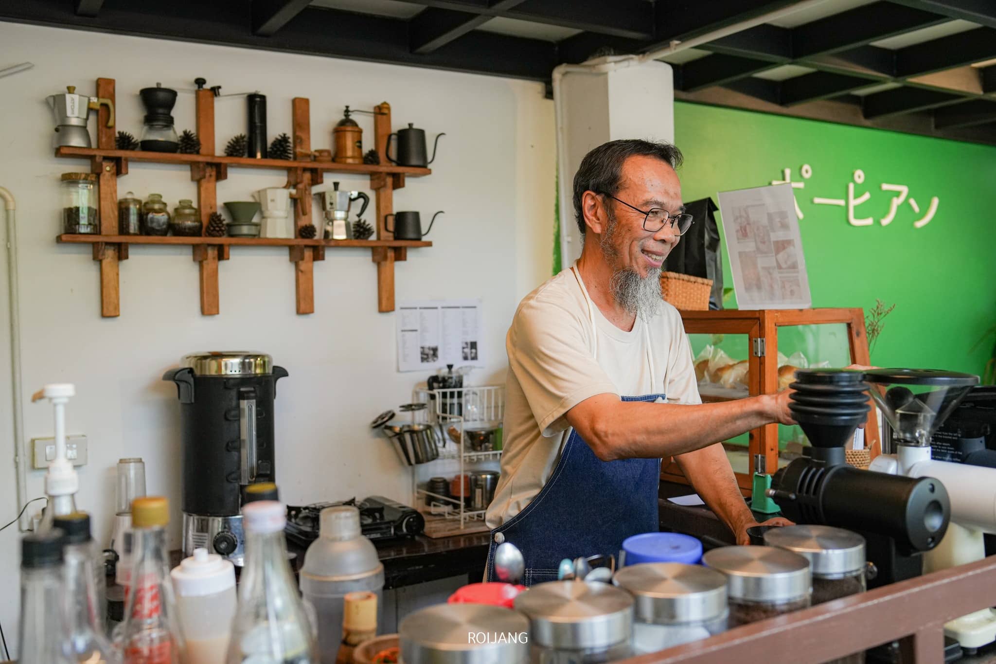 บาริสต้าสวมผ้ากันเปื้อนยืนอยู่หลัง คาเฟ่ภูเก็ต เคาน์เตอร์ร้านกาแฟภูเก็ต ล้อมรอบไปด้วยอุปกรณ์และขวดต่างๆ กำลังเตรียมเครื่องดื่มอย่างพิถีพิถัน ชั้นวางของที่ประดับด้วยอุปกรณ์ชงกาแฟเรียงรายอยู่ตามผนัง ขณะที่ผนังสีเขียวที่มีข้อความเป็นฉากหลังอันสวยงาม.