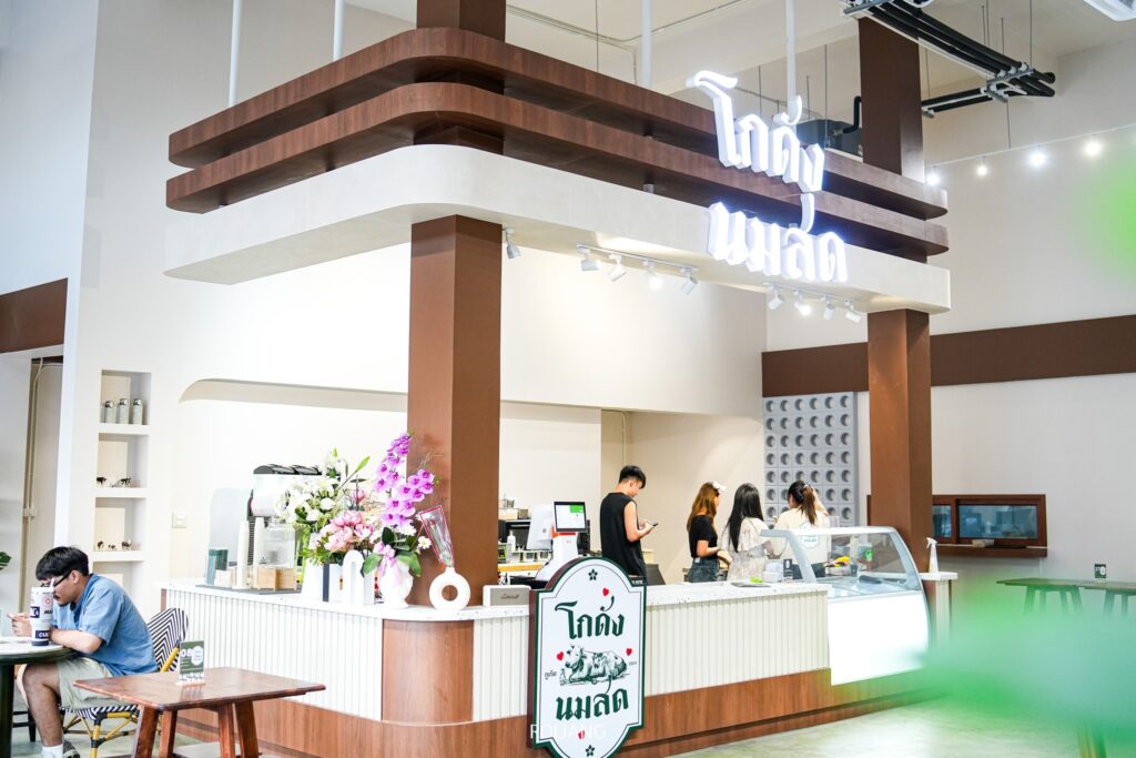 ภายในคาเฟ่ภูเก็ต โกดัง นมสด สไตล์โมเดิร์น ผู้คนต่างสั่งอาหารกันที่เคาน์เตอร์ใต้ป้ายนีออน มีผู้ชายคนหนึ่งนั่งอยู่ที่โต๊ะทางซ้ายมือ การตกแต่งร้านเป็นแบบร่วมสมัยด้วยไม้และองค์ประกอบสีขาว.