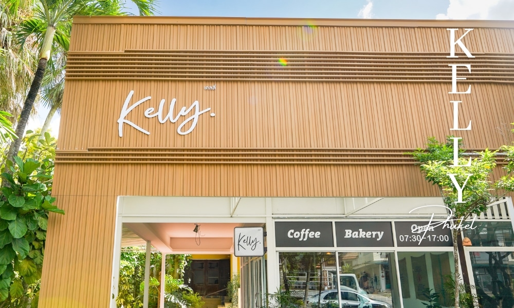 มุมมองภายนอกของร้านกาแฟ "Kelly" ร้านกาแฟภูเก็ตที่มีส่วนหน้าอาคารไม้ ร้านกาแฟภูเก็ต  ต้นไม้สีเขียว และป้ายแสดง "กาแฟ" "เบเกอรี่" และเวลาทำการตั้งแต่ 07.30 น. ถึง 17.00 น.