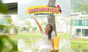 ผู้หญิงคนหนึ่งนั่งยิ้มอยู่บนเก้าอี้สีเหลืองนอกร้าน Rose River Café คาเฟ่ภูเก็ตอันมีเสน่ห์ ซึ่งมีสระน้ำอันเงียบสงบและอาคารต่างๆ เป็นฉากหลัง.