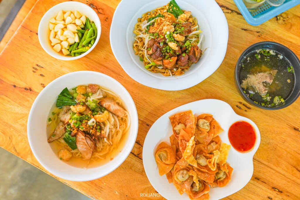 ที่ บะหมี่พังงาตลาดขวาง เชิญคุณเพลิดเพลินกับก๋วยเตี๋ยว 3 ชามพร้อมเครื่องเคียง ชามกระเทียมพริก ซุปใสเนื้อนุ่ม และจานเกี๊ยวทอดพร้อมน้ำจิ้ม จัดเรียงอย่างสวยงามบนโต๊ะไม้สไตล์ชนบท.