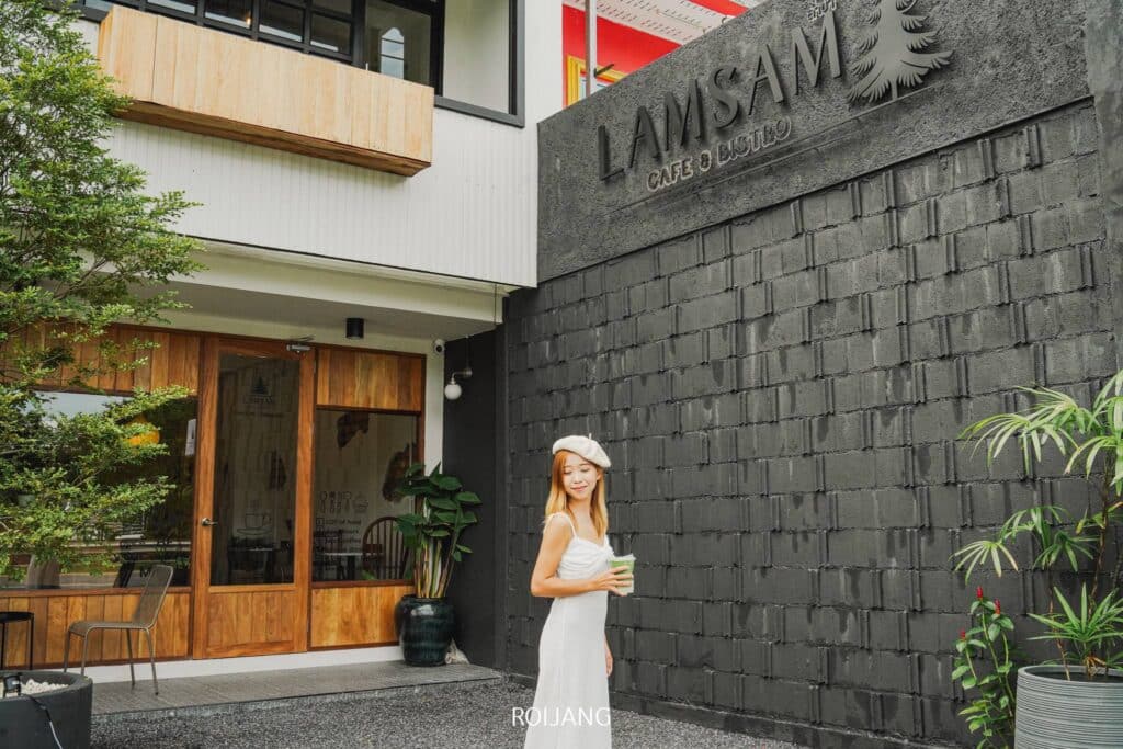 ผู้หญิงสวมชุดสีขาวและหมวกเบเร่ต์ยืนดื่มเครื่องดื่มอยู่หน้า Lamsam Café & Bistro คาเฟ่เขาหลัก ซึ่งภายนอกเป็นอิฐสีดำและประตูไม้ คาเฟ่เขาหลัก