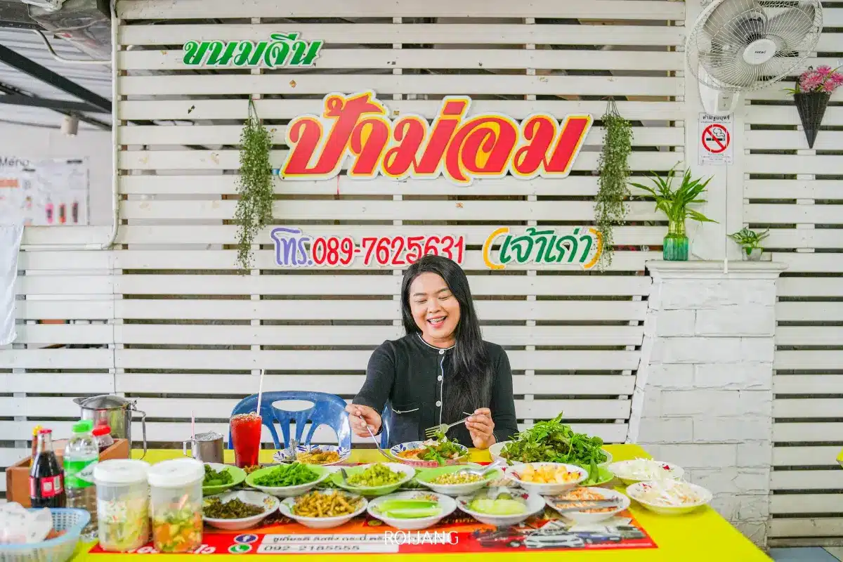 ผู้หญิงคนหนึ่งนั่งอยู่ที่โต๊ะซึ่งเต็มไปด้วยอาหารไทยหลากหลายชนิด เธอยิ้มอย่างมีความสุขขณะรับประทานขนมจีนพังงา โดยมีป้ายชื่อร้านอาหารและหมายเลขโทรศัพท์ติดอยู่บนผนังด้านหลัง.