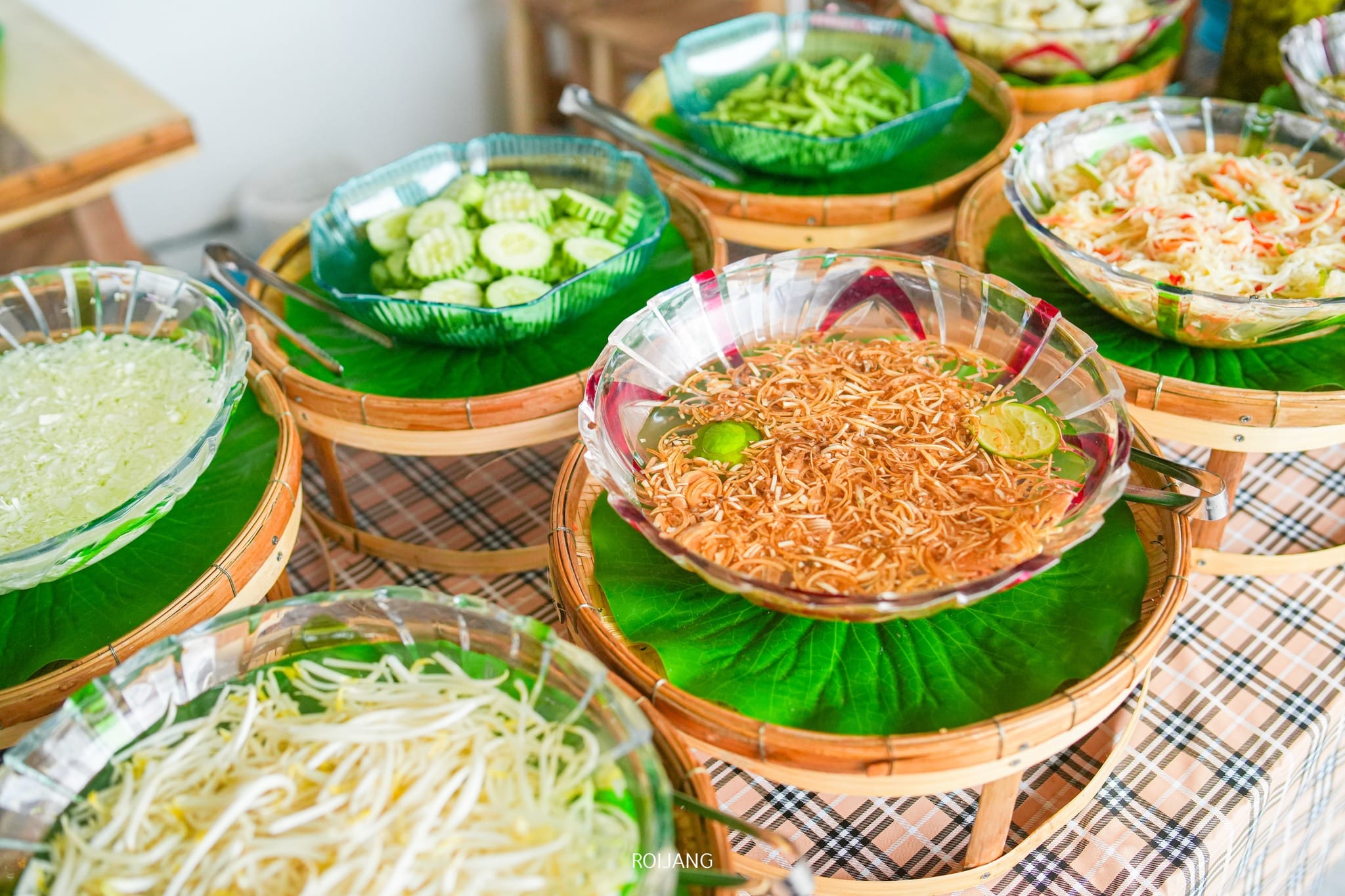 สลัดและผักหั่น ร้านขนมจีนภูเก็ต บางประเภท รวมถึงขนมจีนภูเก็ตแท้ๆ ในชามใสที่แสดงบนใบไม้สีเขียวบนแผงไม้