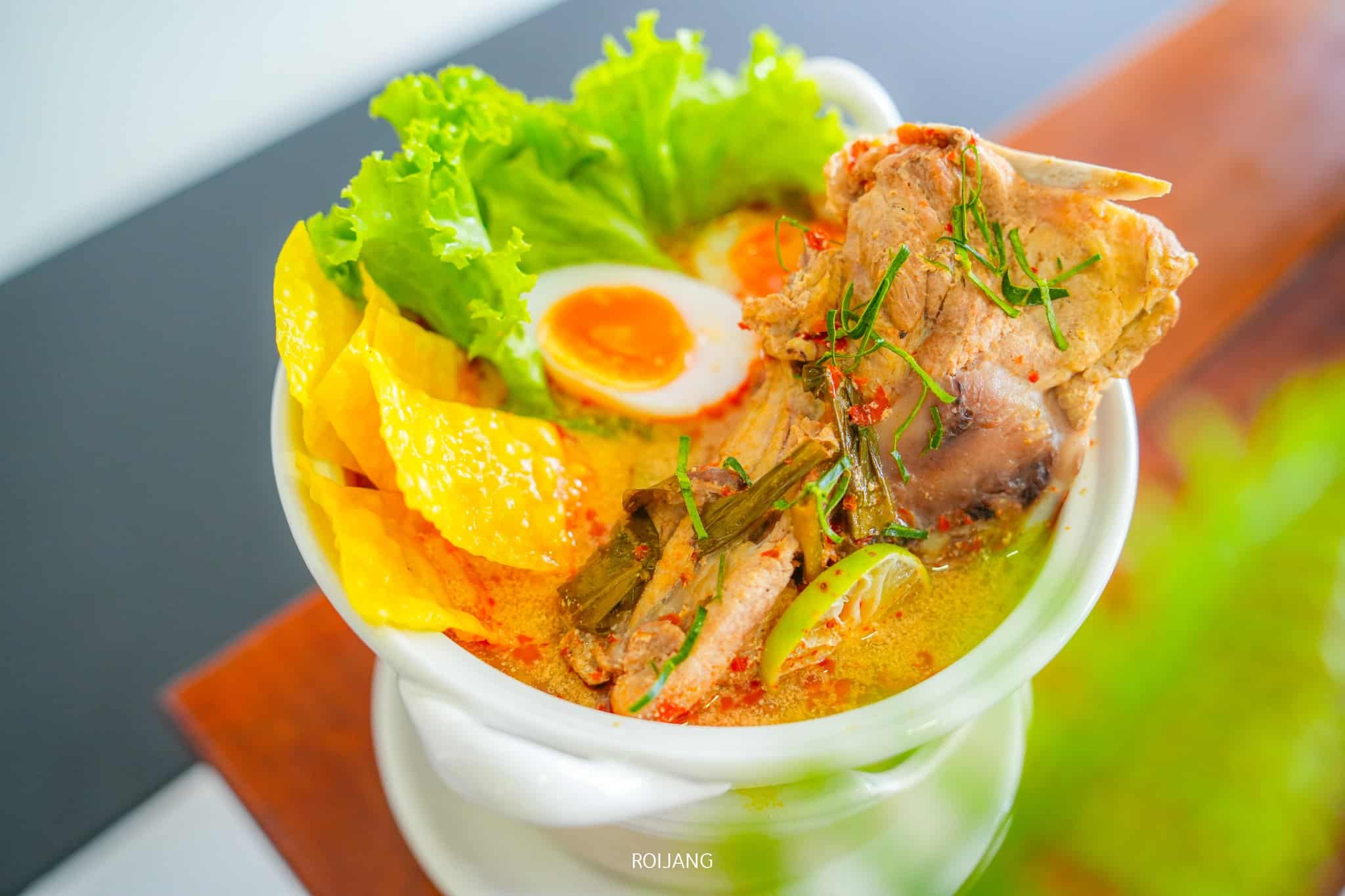 ชามซุปก๋วยเตี๋ยว ร้านอาหารภูเก็ต พร้อมเนื้อสไลซ์ ไข่ต้มผ่าครึ่ง เกี๊ยวสีเหลือง และผักใบเขียว โรยหน้าด้วยสมุนไพรในชามสีขาวบนโต๊ะไม้ที่ชาบูคอง ภูเก็ต ค้นพบอาหารจานเด็ดนี้ที่ร้านอาหารภูเก็ตอันเลื่องชื่อ.